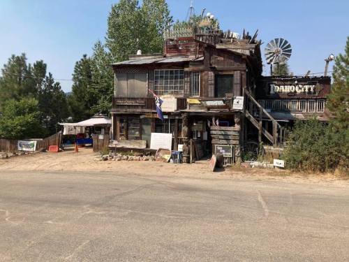 A rustic, weathered building with a windmill, surrounded by trees and a dirt road, featuring various signs and decorations.