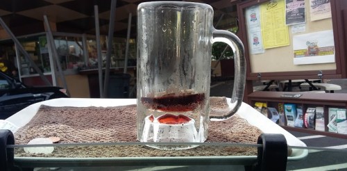 A frosty glass mug filled with dark soda sits on a tray, with a textured surface and a restaurant backdrop.