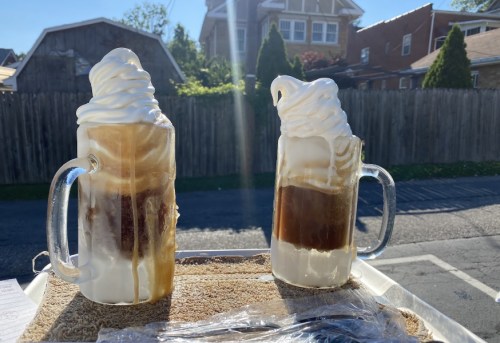 Two frosty mugs of root beer floats topped with whipped cream, set against a sunny outdoor background.