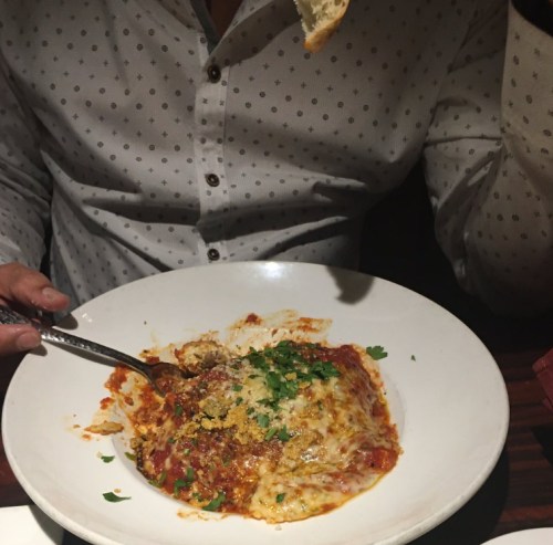 A person holds a piece of bread while enjoying a plate of cheesy lasagna topped with herbs and breadcrumbs.