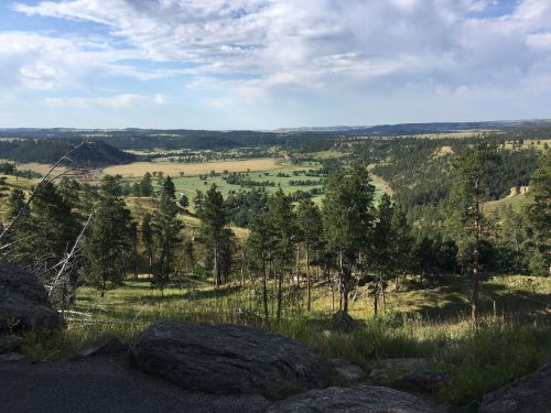 A scenic view of rolling hills and green valleys, framed by trees under a partly cloudy sky.