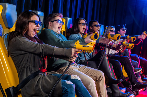 A group of six people wearing 3D glasses excitedly aim toy guns while seated on a ride.