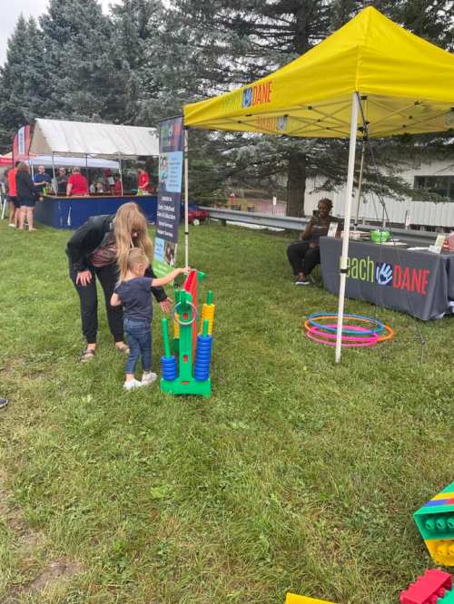 A child plays with colorful toys at an outdoor event, while an adult assists nearby under a yellow tent.