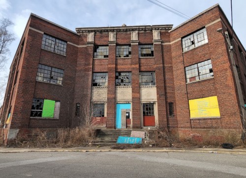Abandoned brick building with broken windows and colorful graffiti on the facade, surrounded by overgrown vegetation.