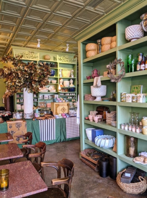 A cozy café interior featuring green shelves filled with pottery, plants, and decorative items, with wooden tables in the foreground.
