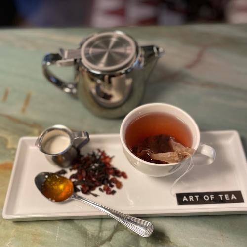 A silver teapot beside a cup of tea with a teabag, a small creamer, dried tea leaves, and honey on a white tray.