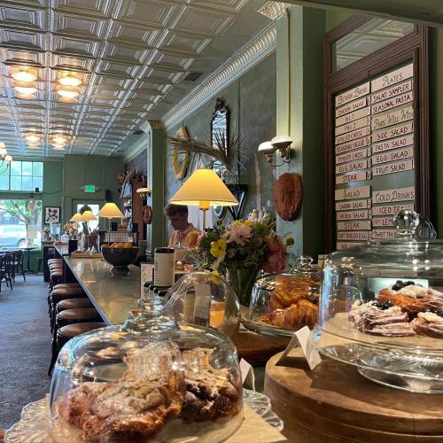 A cozy café interior with a counter displaying pastries, flowers, and a menu on the wall. Soft lighting creates a warm atmosphere.