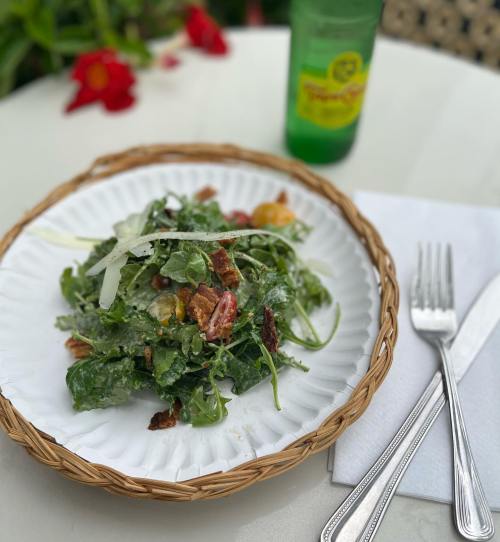 A fresh salad with greens, nuts, and cheese on a woven plate, accompanied by a green beverage and silverware.