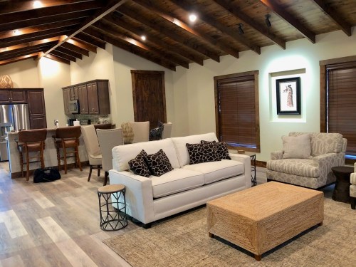 Cozy living room with a white sofa, patterned chairs, wooden beams, and a kitchen in the background.