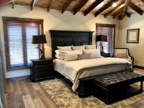 Cozy bedroom featuring a dark wood bed, nightstands, lamps, and large windows with blinds. Rustic decor and warm tones.