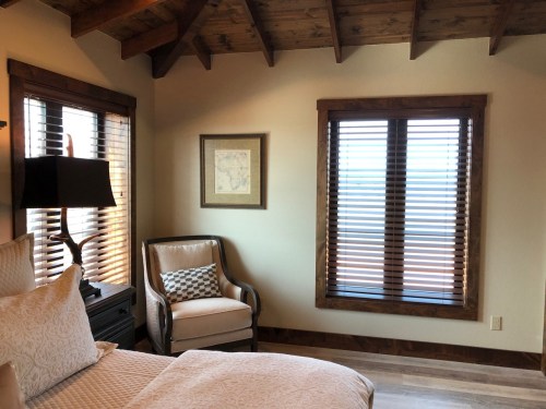 Cozy bedroom with wooden beams, a bed, a chair, and windows with blinds, creating a warm, inviting atmosphere.