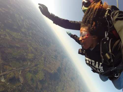 A person skydives, arms outstretched, with a scenic landscape below and another skydiver above them.