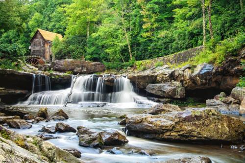 A serene waterfall cascades over rocks near a rustic wooden mill, surrounded by lush green trees.
