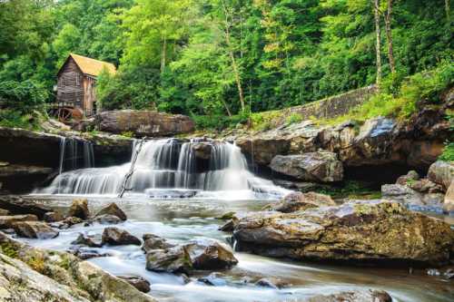 A serene waterfall cascades over rocks near a rustic wooden mill, surrounded by lush green trees.