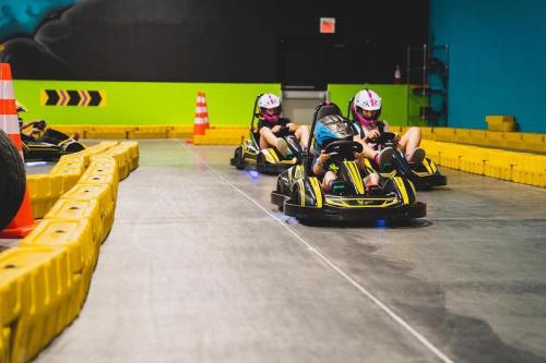 Two go-karts racing on an indoor track, with bright yellow barriers and traffic cones in the background.