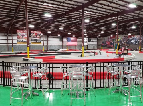 Indoor go-kart track with a circular layout, surrounded by barriers and seating, featuring an American flag on the wall.