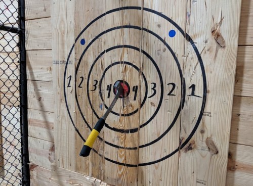 A wooden target with concentric circles, featuring a knife embedded in the center, surrounded by scoring numbers.