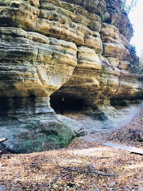 A person stands in front of a large, textured rock formation with layered cliffs and a dirt path in a wooded area.