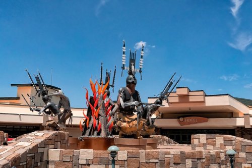 A large sculpture of warriors with spears and flames, set against a blue sky near a hotel entrance.