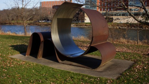 A large, abstract metal sculpture with curved shapes, set in a grassy area near a river.