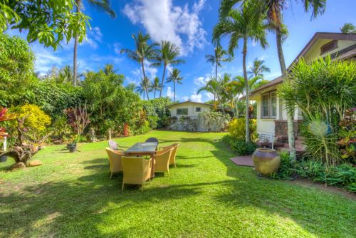 A lush garden with palm trees, a seating area, and a house under a bright blue sky.
