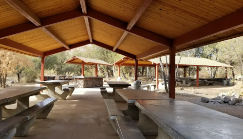 A spacious outdoor picnic area with covered tables and a stone grill, surrounded by trees and natural scenery.
