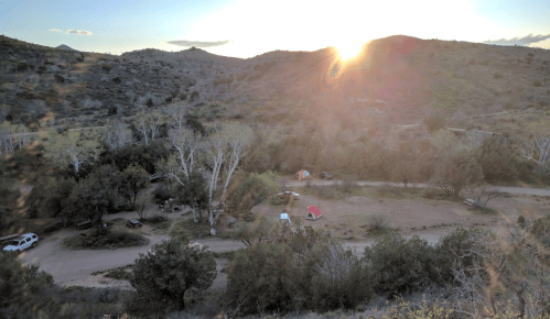 A scenic view of a campsite in a valley at sunset, with tents and trees surrounded by rolling hills.