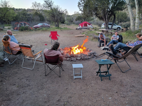 A group of people sitting around a campfire in a campsite, with tents and trees in the background.
