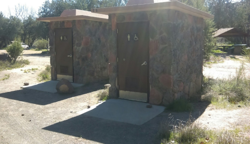 Two stone restroom buildings with accessible signs, set in a natural outdoor area with sparse vegetation.