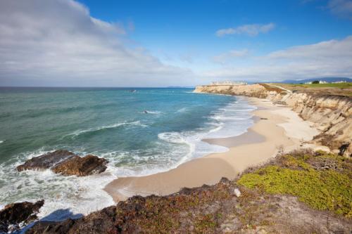 A scenic coastal view with sandy beach, rocky shoreline, and gentle waves under a partly cloudy sky.