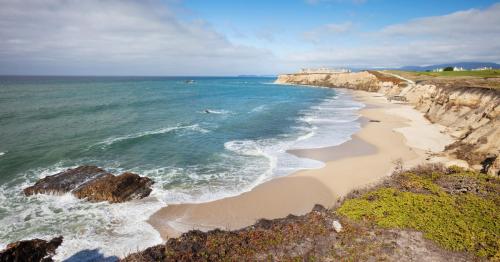 A scenic view of a sandy beach with rocky cliffs and gentle waves under a partly cloudy sky.