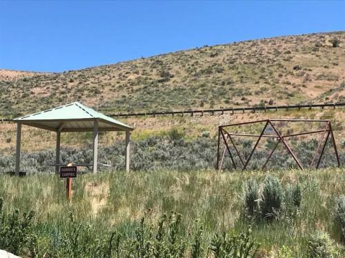 A small gazebo in a grassy area with a metal structure nearby, set against a hilly landscape under a clear blue sky.
