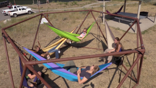 Five people relax in colorful hammocks within a metal frame structure on a grassy area.