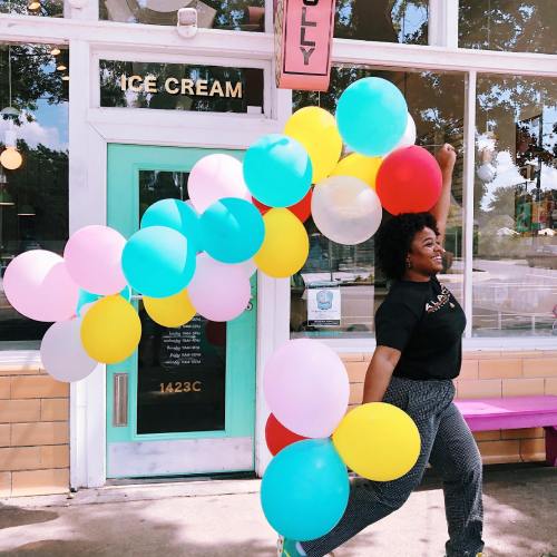 A joyful person poses with colorful balloons outside an ice cream shop, showcasing a vibrant and festive atmosphere.