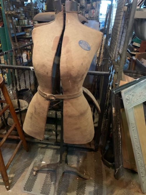 A vintage dress form with a worn fabric cover, displayed in a rustic shop surrounded by various items.