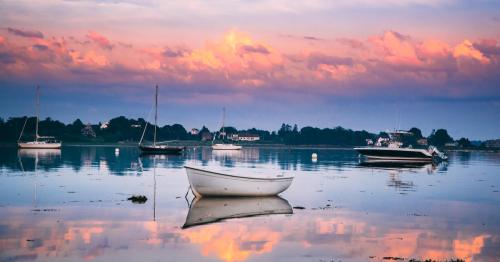 A serene lake at sunset with a small white boat and several sailboats reflected in the calm water.