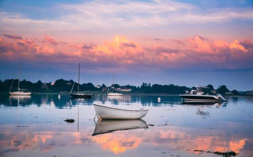 A serene water scene at sunset with a small white boat and colorful clouds reflecting on the calm surface.