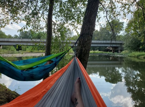 Two hammocks are set up between trees by a calm river, with a bridge visible in the background and greenery surrounding the scene.