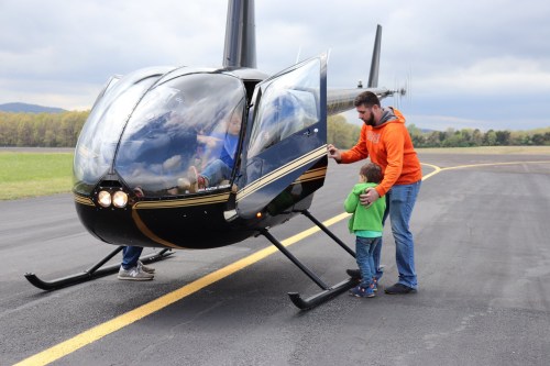A man and a child approach a helicopter on a runway, with the helicopter's door open and passengers inside.