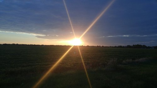 A sunset over a green field, with rays of light shining through clouds in a blue sky.