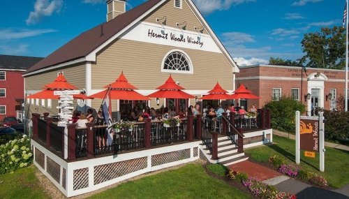 A winery with outdoor seating under orange umbrellas, surrounded by greenery and a blue sky.