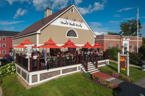 A restaurant with outdoor seating under red umbrellas, surrounded by greenery and a blue sky with clouds.