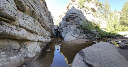 A serene rocky landscape with a small waterfall and calm water reflecting the surrounding cliffs and trees.