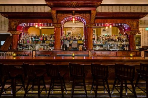 A beautifully designed bar with ornate woodwork, shelves lined with bottles, and empty stools in a warm, inviting setting.
