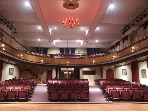Interior of a theater featuring rows of red seats, a stage, and a chandelier, with a balcony above.