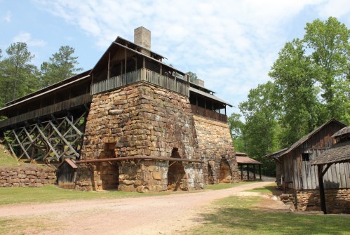 Historic stone furnace building with wooden structures nearby, surrounded by greenery and a clear blue sky.