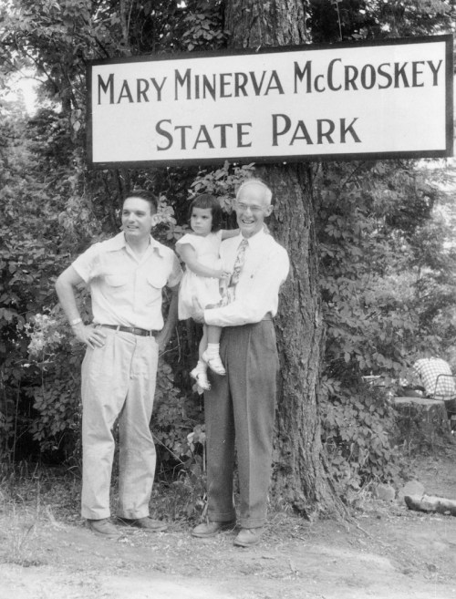 Two men and a young girl pose in front of a sign for Mary Minerva McCroskey State Park, surrounded by trees.