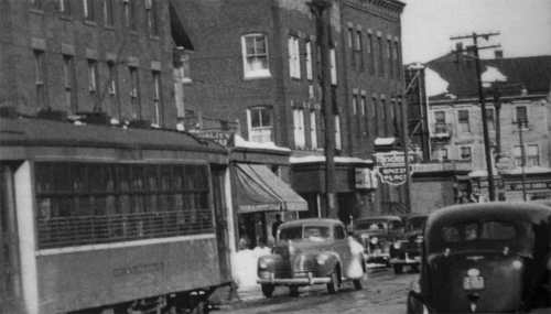 Black and white image of a busy street with vintage cars and a streetcar, featuring storefronts and buildings.