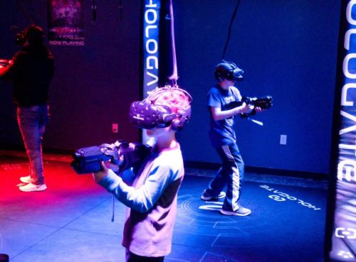 Two children wearing VR headsets hold controllers in a dimly lit room with colorful lights.
