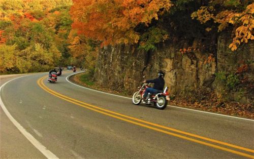 Motorcyclists riding along a winding road surrounded by vibrant autumn foliage and rocky cliffs.